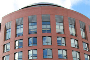 A red brick building with a cylindrical shape, featuring multiple windows in a modern architectural style against a partly cloudy sky.