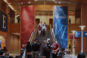 People walking and sitting in an indoor area of an academic building, with large banners displaying "Wharton" and other text hanging prominently.