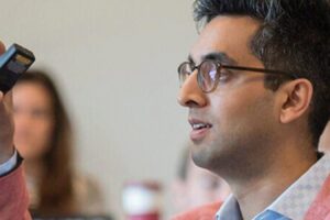 A person in glasses and a pink jacket is engaged in a discussion or presentation, gesturing with a remote. Several people in the background are listening.